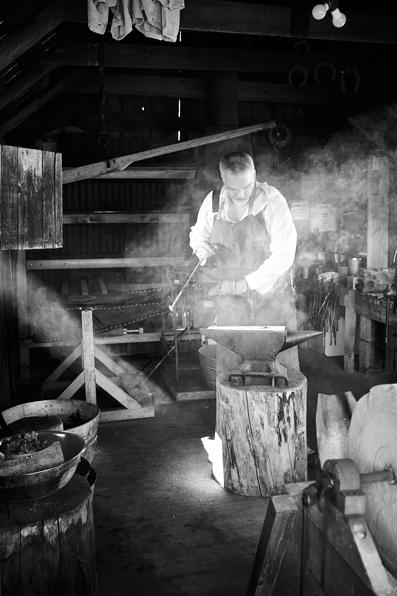 Fujifilm X100V monochrome SDR JPEG of a blacksmith working beside a bright window.