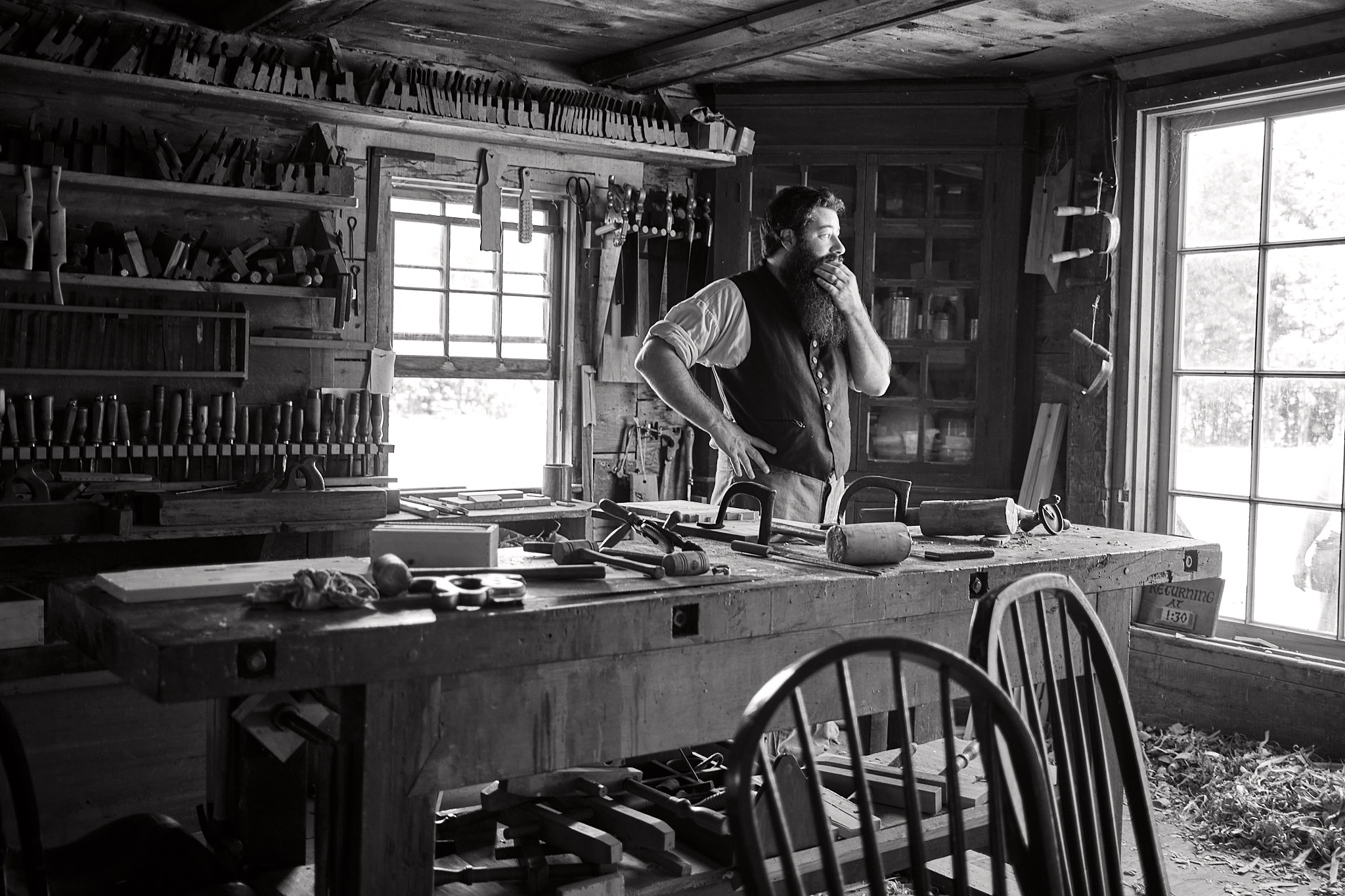 Fujifilm X100V SOOC Acros JPEG of a bearded craftsperson standing in a dark woodshop beside bright windows and benches full of tools.